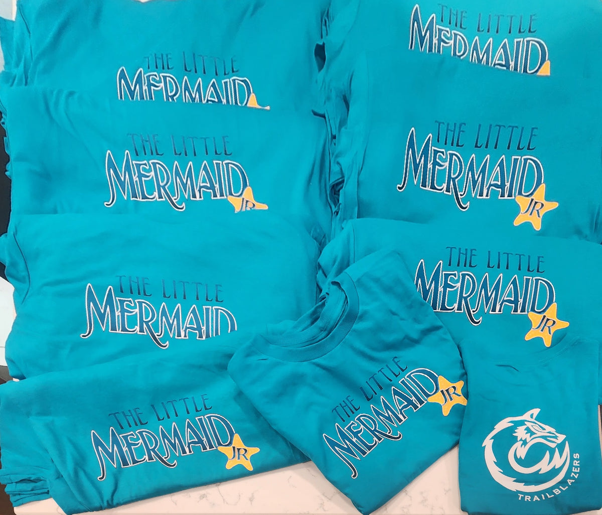 A stack of turquoise blue t-shirts featuring 'The Little Mermaid Jr.' graphic prints in white and yellow, with a Trailblazer logo on one shirt.